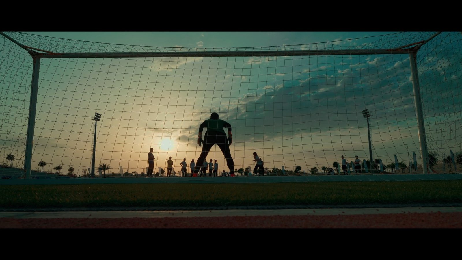 Anamorphic documentary still: a goalkeeper silhouetted against a sunset behind the goal net.