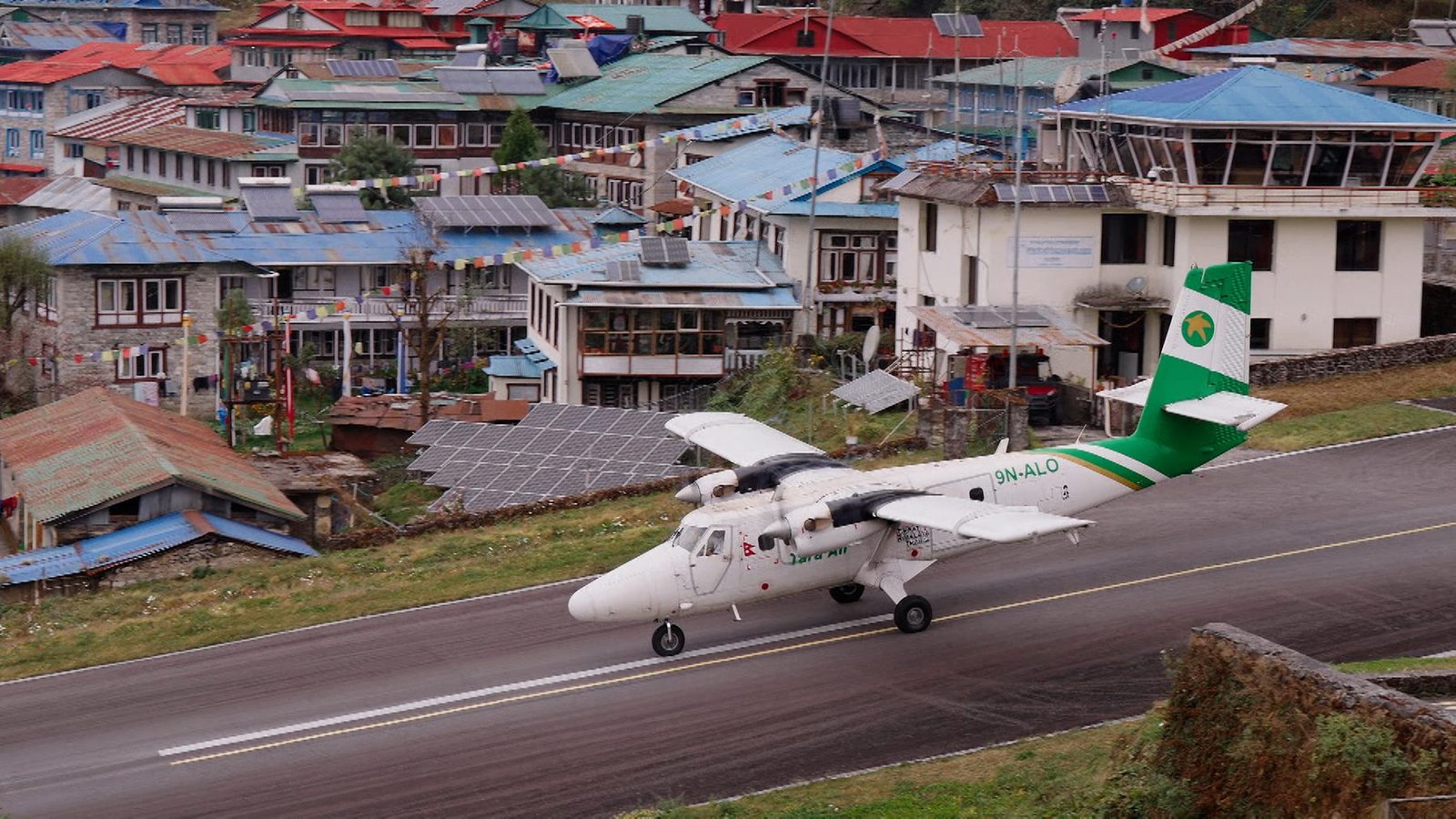TV series still: a Twin Otter aircraft landing at Lukla, gateway to the Everest region, colourful Himalayan village behind.